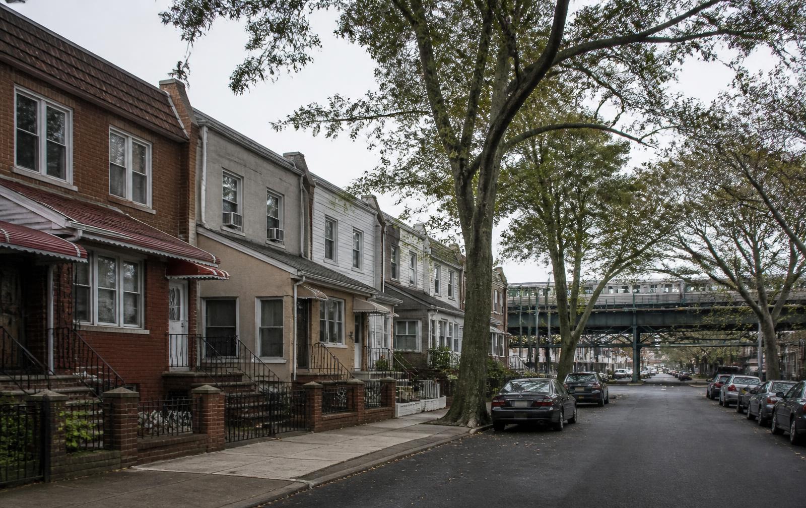 Quiet residential street of brick row houses in Queens, New York with an elevated train trestle beyond